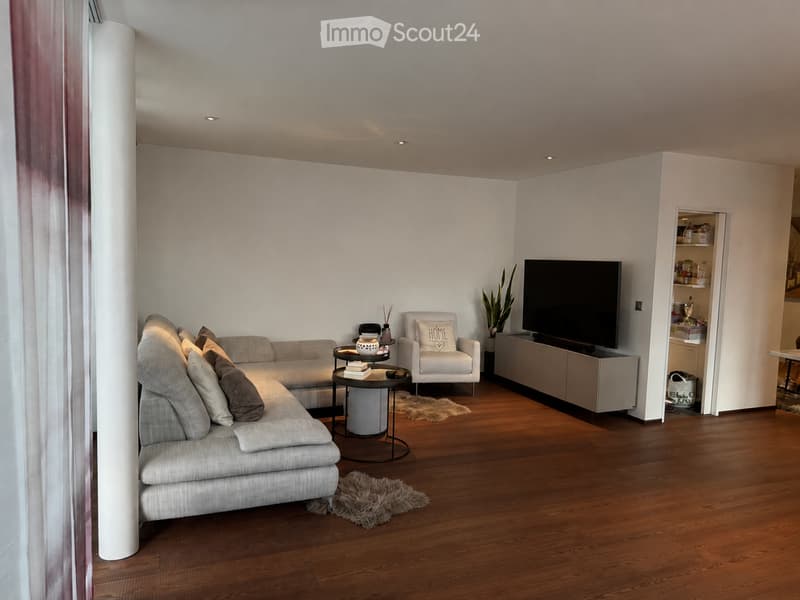 Modern living room with a grey sectional sofa, a white armchair, a coffee table, a flat-screen TV, and a small plant. The room features wooden flooring and white walls.