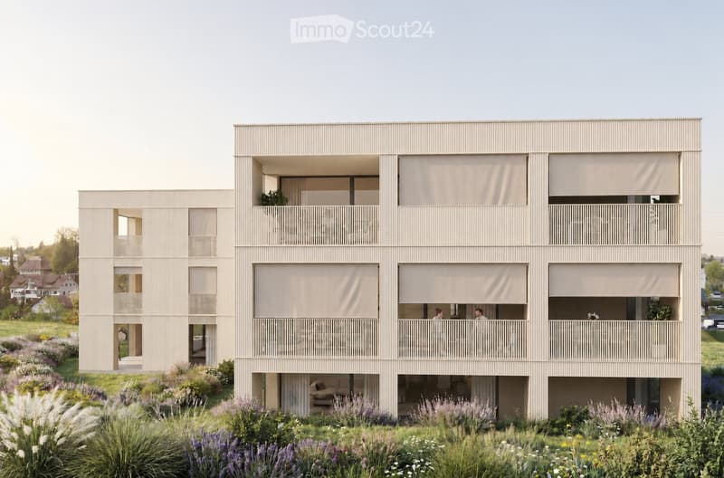 Modern two-story building with beige facade, large windows, and balconies. The ground floor has a raised entrance, and the building is surrounded by a well-maintained garden with various plants and flowers.