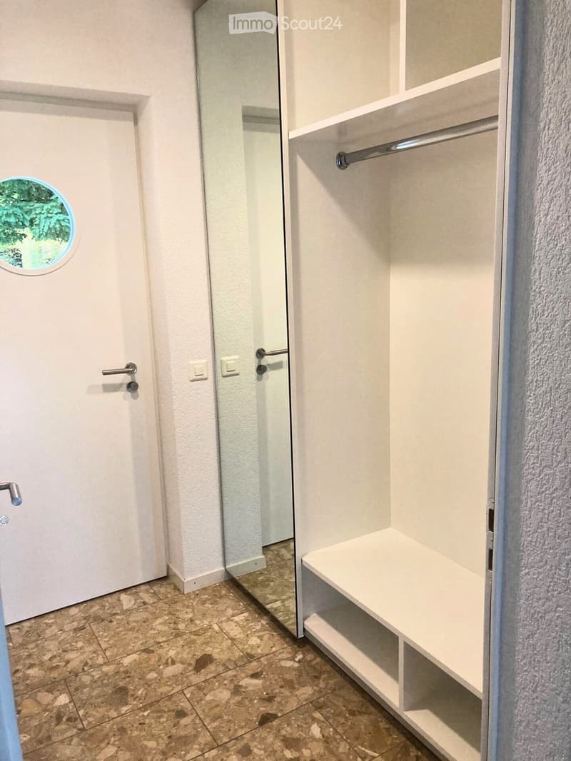 Empty hallway with tiled floor, white walls, and a built-in closet with shelves and a hanging rod. A round window with a view of greenery is visible on the left.
