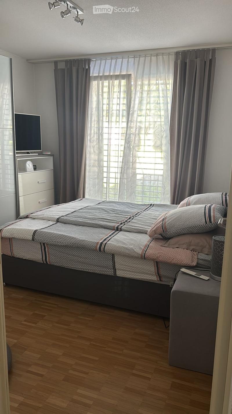A modern bedroom with a large bed, patterned bedding, nightstand, and a TV on a stand. The room features wooden flooring and large windows with curtains.