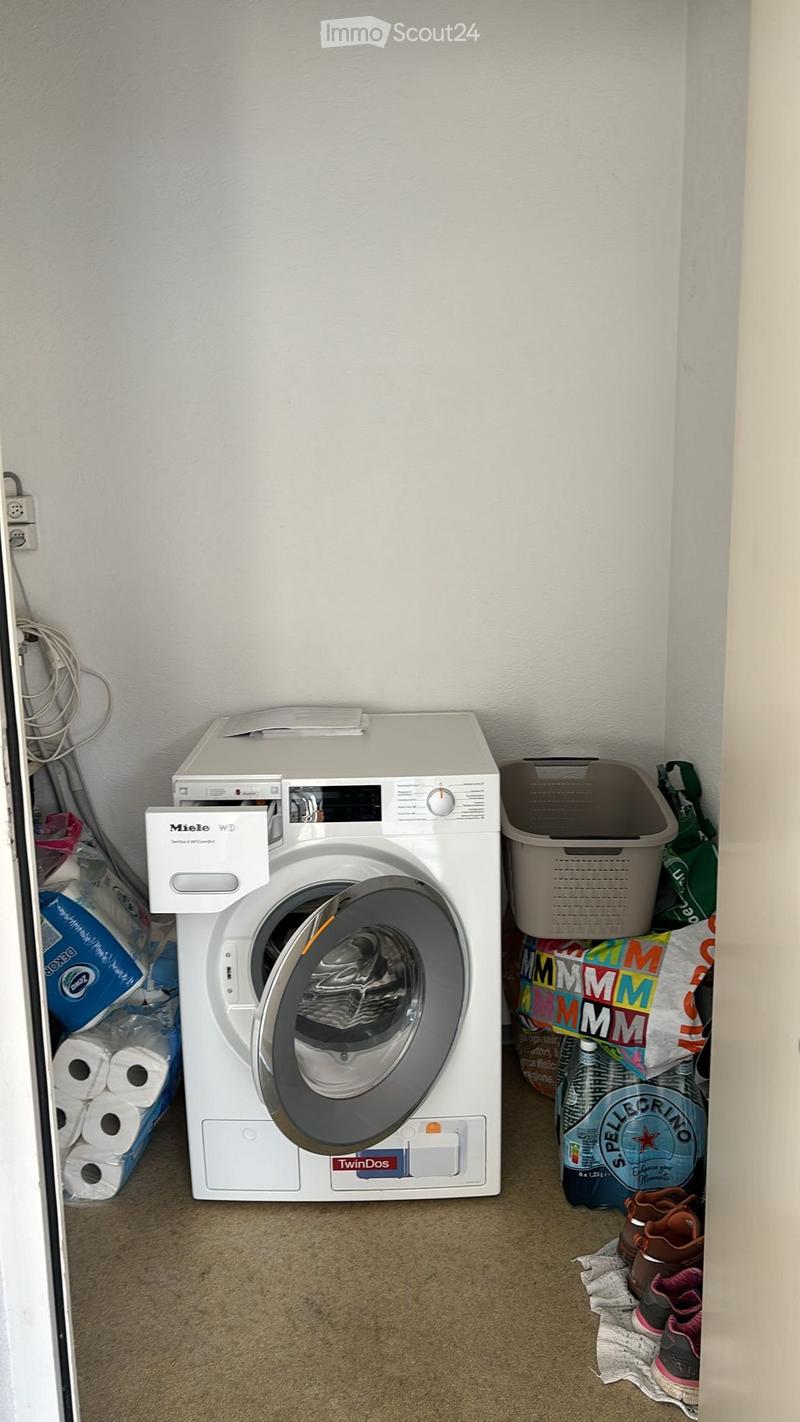 A small laundry area with a white washing machine, a laundry basket, toilet paper, a bottle, and shoes on the floor.