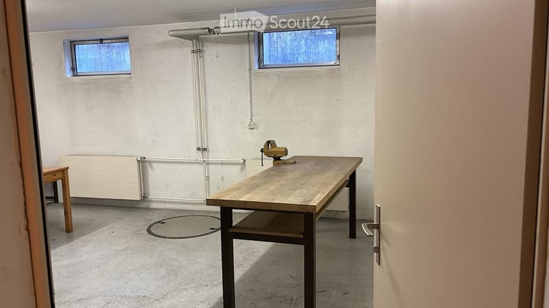 A small, unfurnished room with a wooden table and a stool, white walls, and two windows.