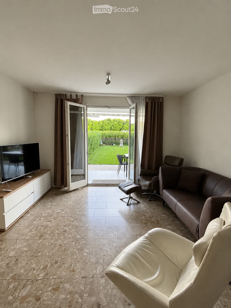 Modern living room with a brown leather sofa, white armchair, and a TV on a white stand. Large sliding glass doors open to a garden view.