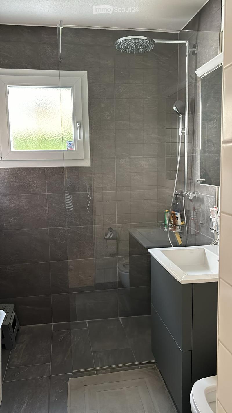 Modern bathroom with dark grey tiles, glass shower enclosure, white sink, and toilet. Features a large showerhead and a window with frosted glass.