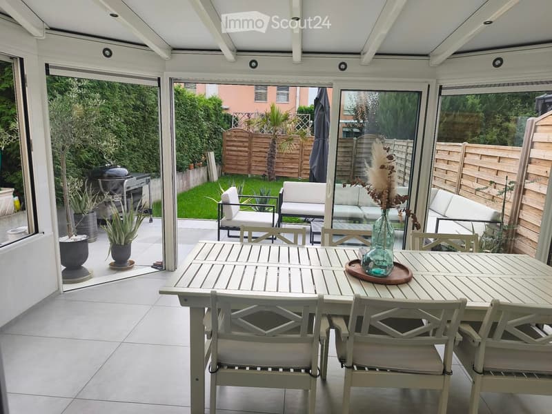 A covered patio with a dining table, chairs, and a vase with dried flowers. The space has large glass doors opening to a garden with a sofa, potted plants, and a barbecue.
