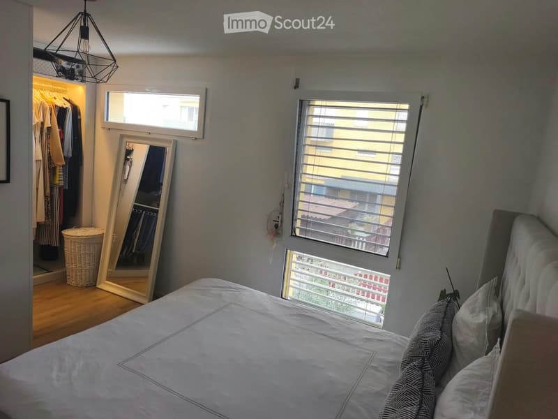 Bedroom with a large bed, a full-length mirror, a wardrobe, and a wicker basket. The room has wooden flooring and large windows with blinds.