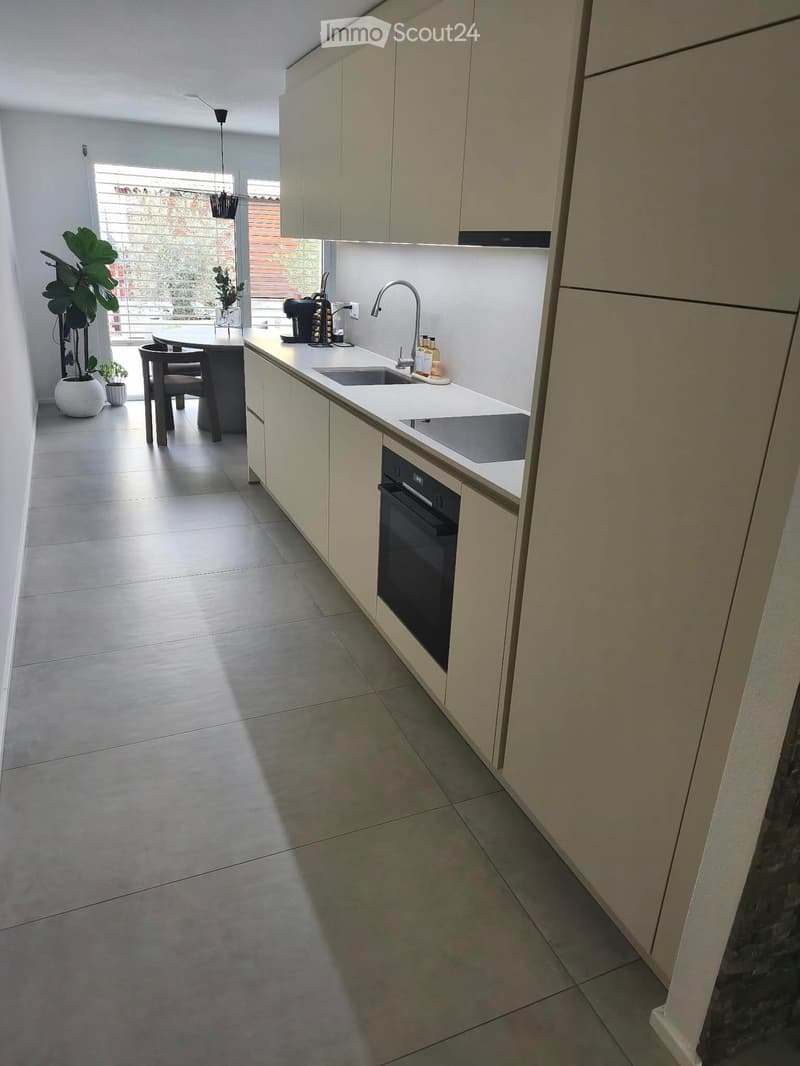 Modern kitchen with white cabinets, stainless steel sink, built-in oven, and a small dining area with a table and chairs. The space features tiled flooring and large windows with blinds.