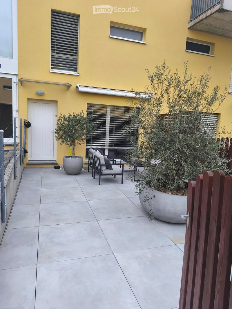 A small outdoor patio area with a tiled floor, a white door leading to the building, two potted olive trees, a lounge chair, and a small table.