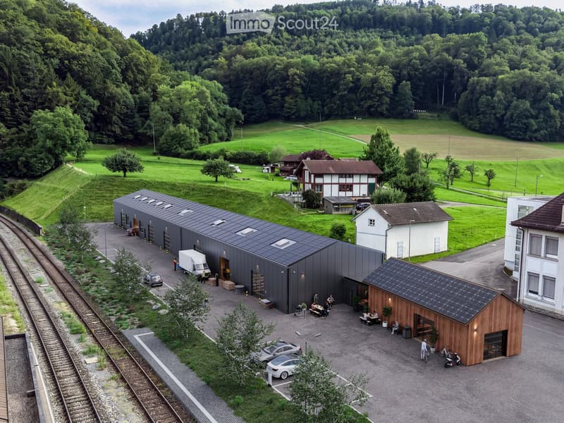 Aerial view of a modern industrial building complex with a large garage, solar panels, and surrounding greenery.
