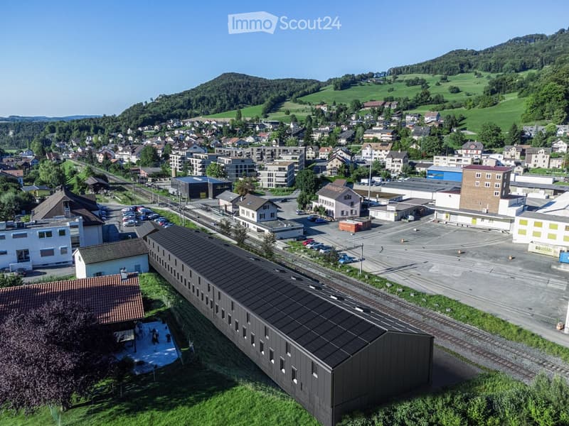 Aerial view of a town with residential and commercial buildings, parking lots, and a train station, surrounded by green hills and scattered trees.