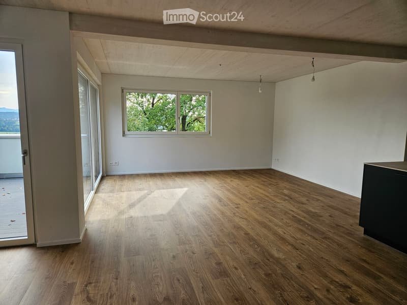 Empty room with wooden flooring, white walls, and large windows allowing natural light. The room features a sliding glass door leading to a balcony or outdoor area.