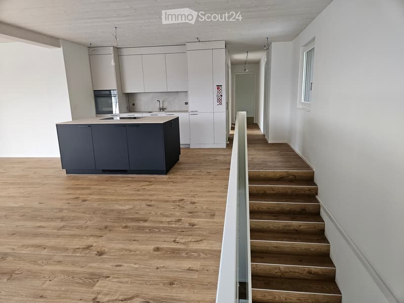 Open-plan space with light wooden flooring, white walls, and a modern kitchen area featuring grey cabinets and a central island. A staircase leads upwards.