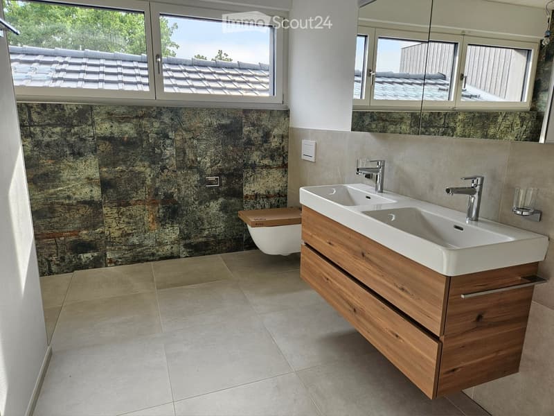 Modern bathroom with a double sink, wooden cabinet, large mirror, and a toilet. The walls feature a stone-like texture, and the floor is tiled.