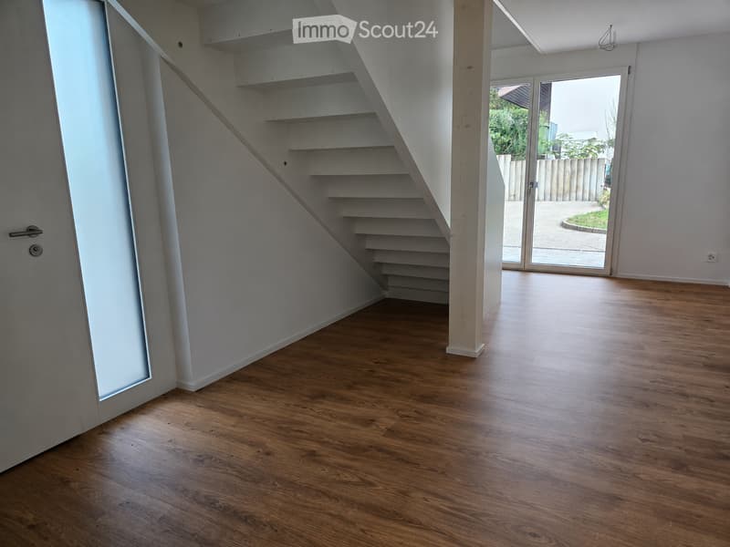 Empty room with wooden flooring, white walls, a staircase leading upwards, and a large glass door opening to an outdoor area.