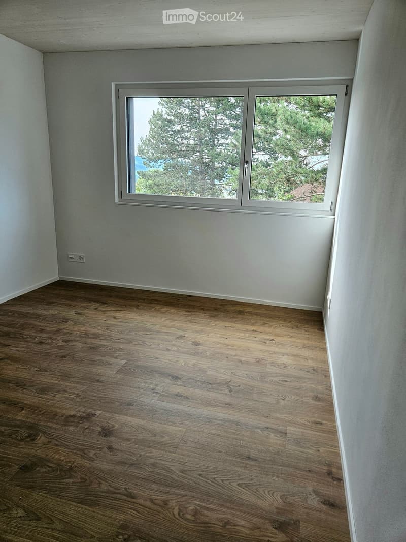 Empty room with wooden flooring and white walls, featuring a large window with a view of trees and a blue sky.