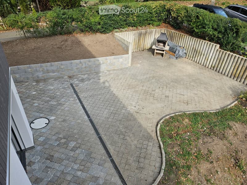 Garden area with a raised concrete border, a small seating area with a table and chairs, and a covered barbecue pit. The ground is paved with stone tiles, and there is a grassy patch and a small raised bed with soil.