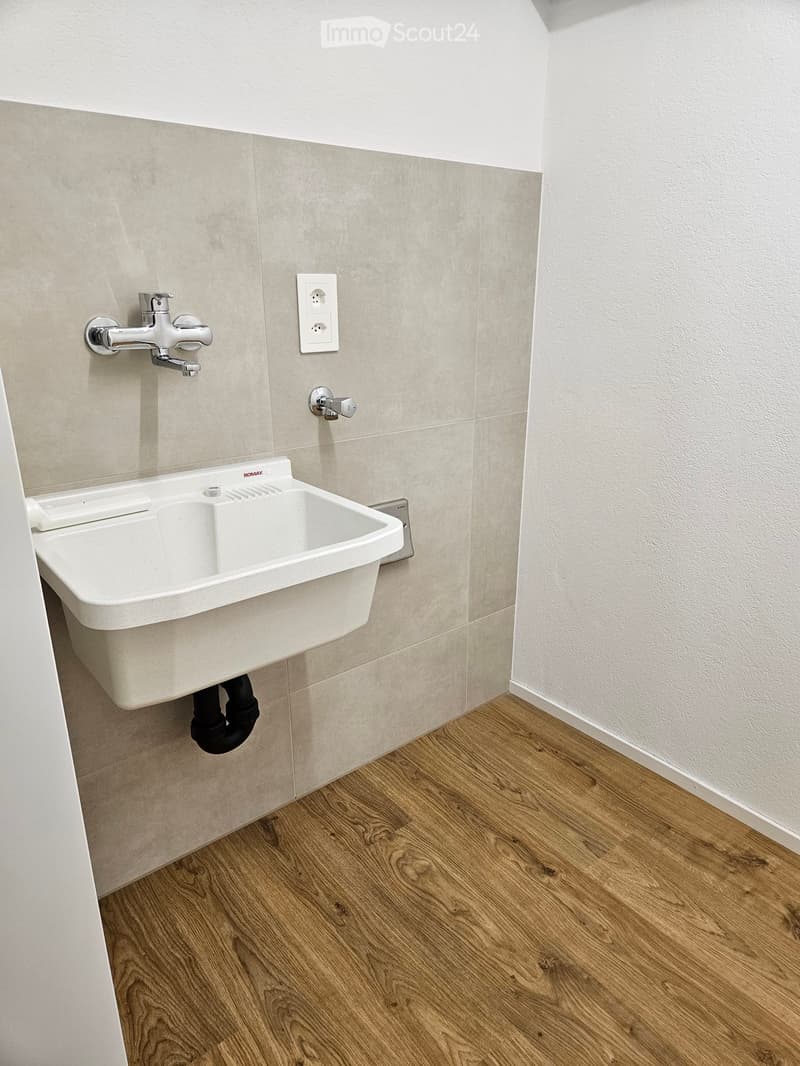 Modern bathroom with a white sink, chrome faucet, and light-colored tiled walls. Wooden flooring is visible.