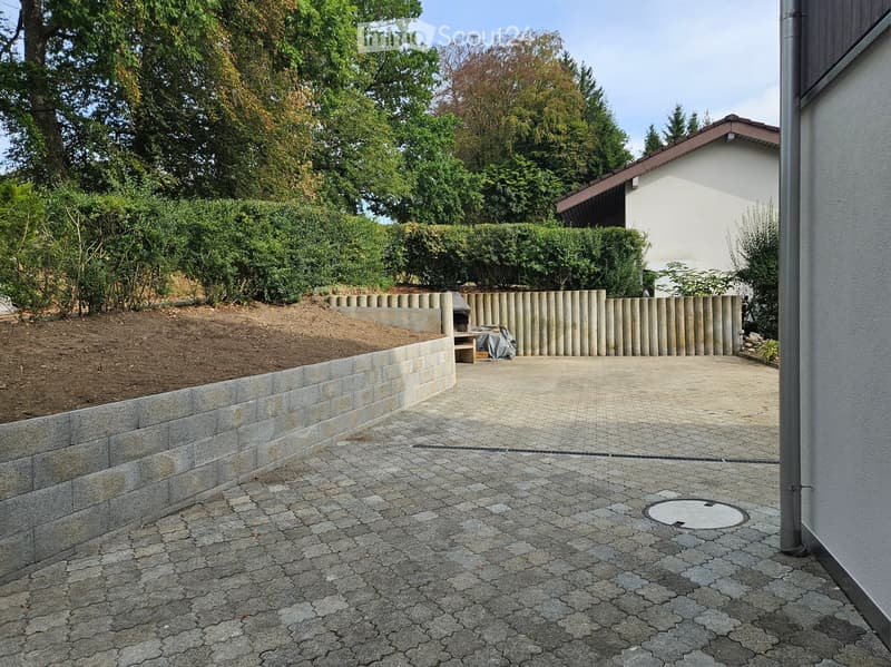 Paved outdoor area with a raised garden bed bordered by a stone wall, surrounded by trees and shrubs, with a covered outdoor seating area and a house in the background.