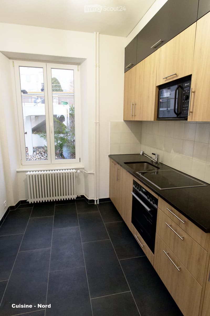 Modern kitchen with dark tiled floor, light wooden cabinets, stainless steel sink, microwave, and oven. Large windows provide natural light and a view of the outdoor area.