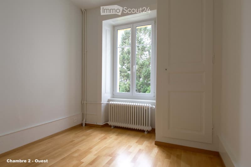 Empty room with wooden flooring, white walls, a large window with white frame, and a white radiator below the window.