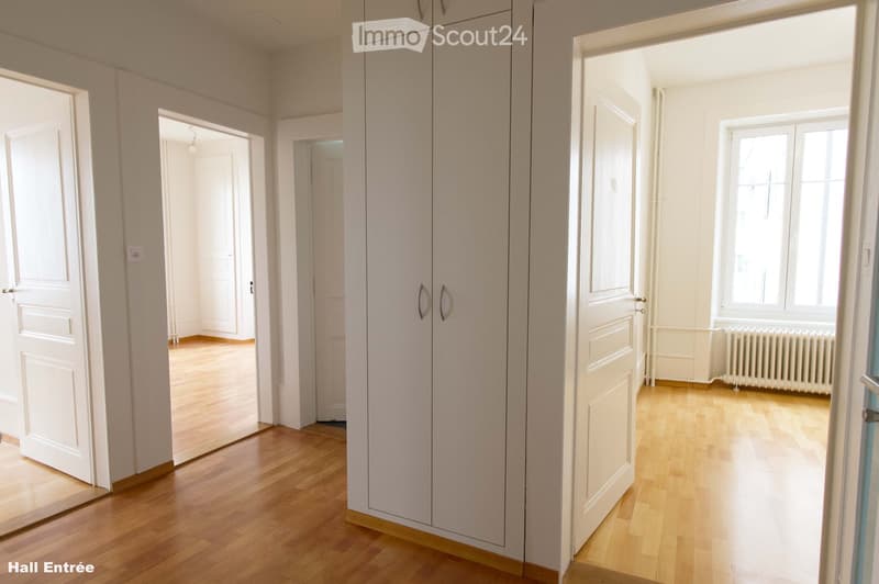 Empty hallway with wooden flooring, white walls, and multiple doors leading to other rooms. Natural light streams in from a window on the right side.