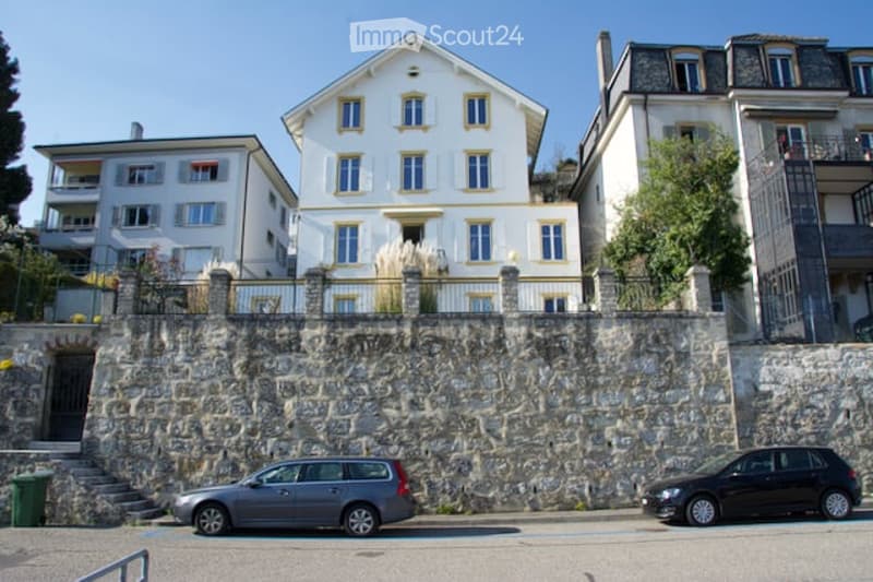 A multi-story residential building with white walls and yellow trim, situated on a raised stone foundation with a small staircase leading to the entrance. Two cars are parked in front of the building.