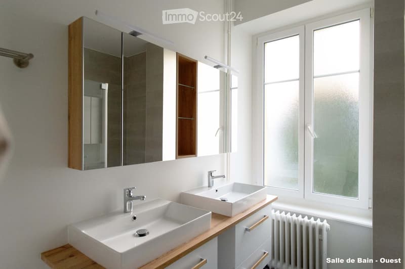 Modern bathroom with two white sinks, chrome faucets, a large mirrored cabinet, and a radiator under a window.