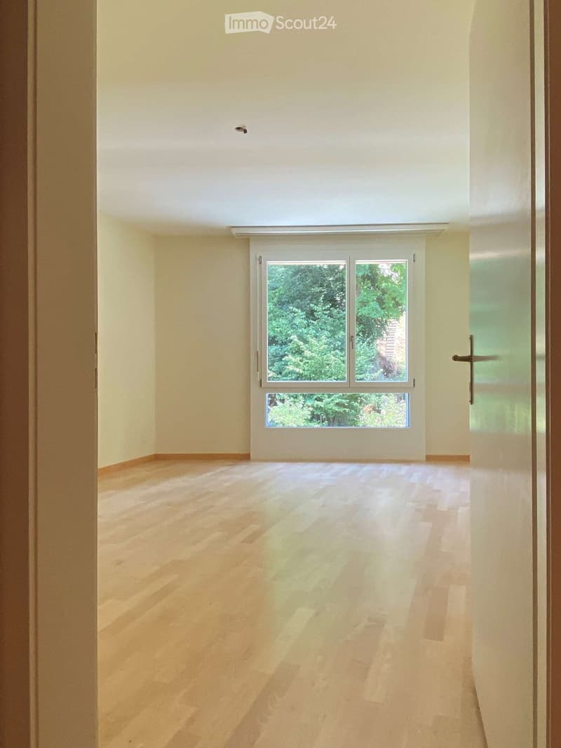 Empty room with light wooden flooring and white walls, featuring a large window with a view of greenery outside.