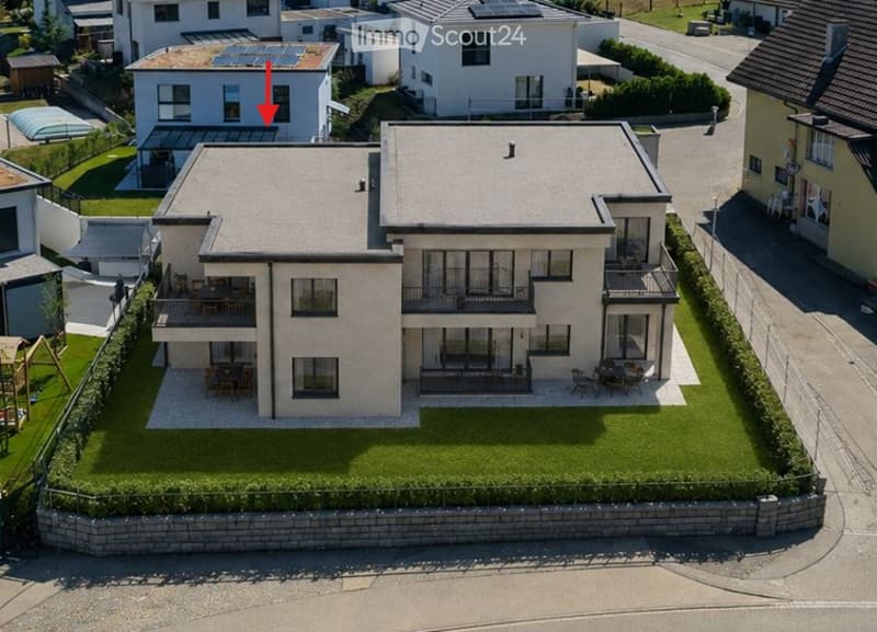 Aerial view of a modern two-story house with a flat roof, surrounded by a well-maintained lawn and neighboring houses. The house features large windows, balconies, and outdoor seating areas.