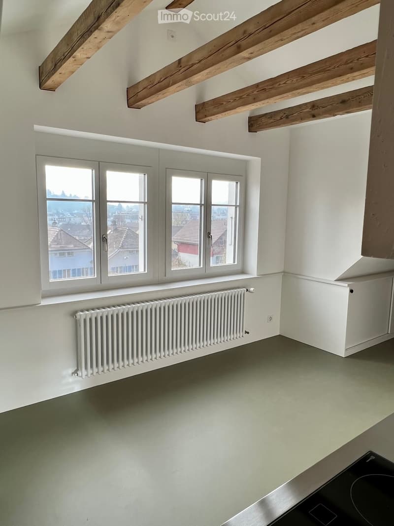 Empty room with exposed wooden beams, large windows offering a view of rooftops, and a radiator under the windows.