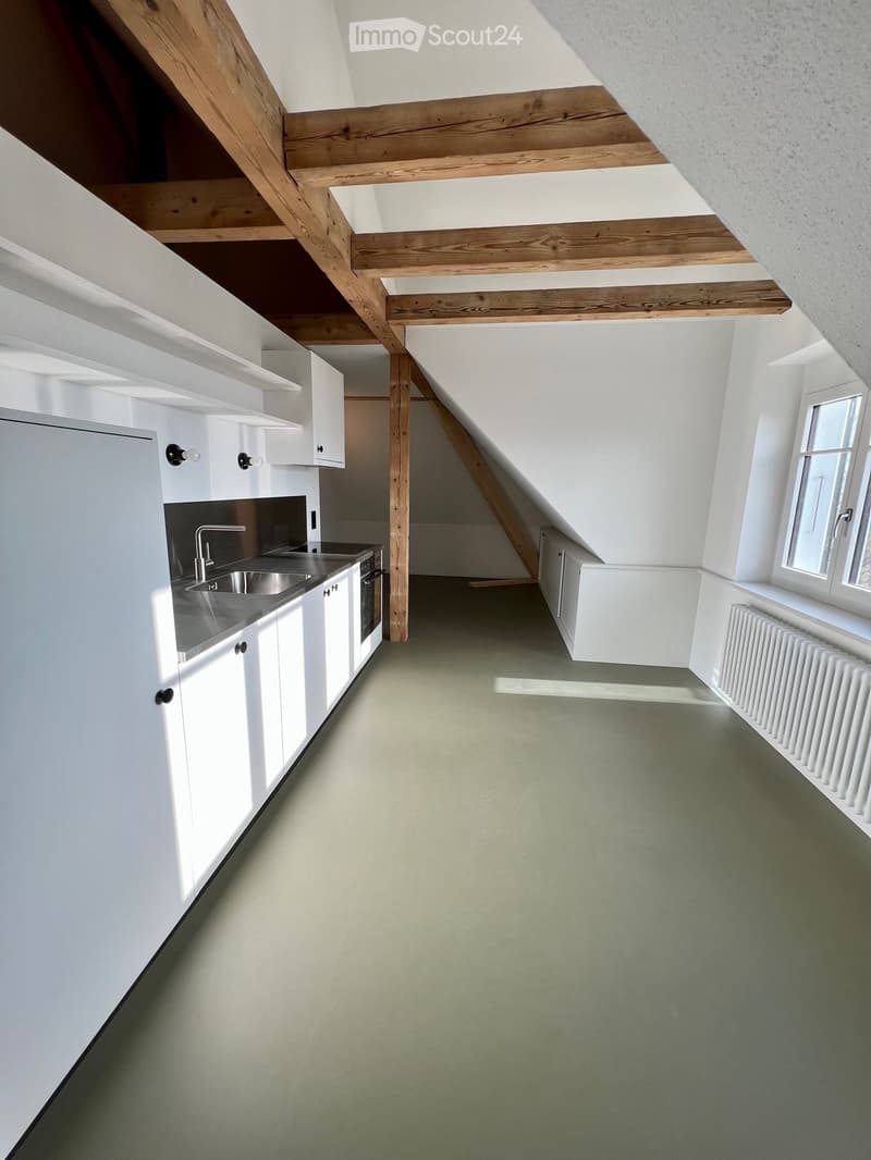 Modern kitchen with white cabinets, stainless steel sink, and exposed wooden beams on the ceiling. Green floor and large window on the right side.