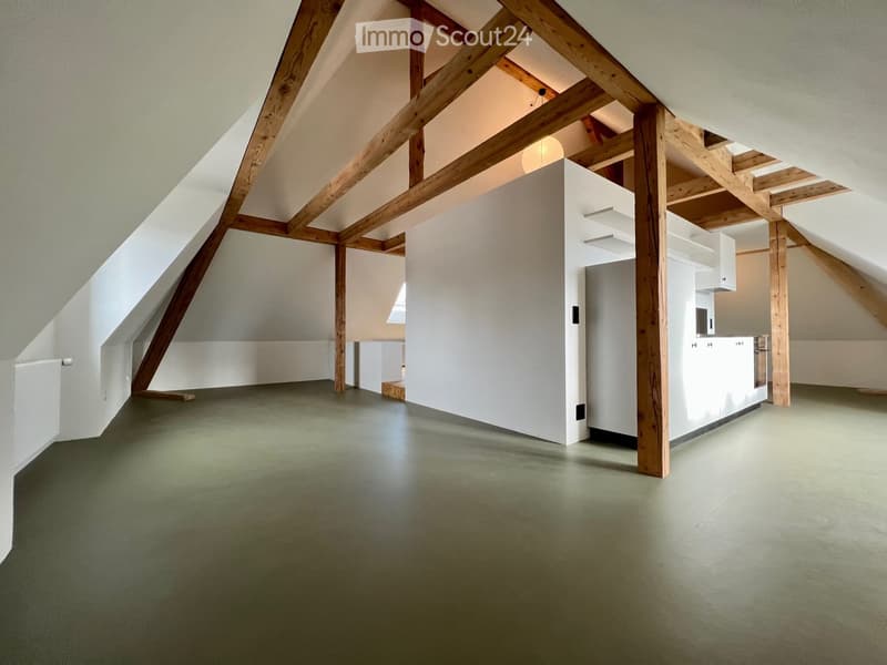 An attic space with exposed wooden beams, white walls, and a modern kitchen area in the corner. The floor is painted green, and the room is well-lit with natural light coming from the sloped ceiling windows.