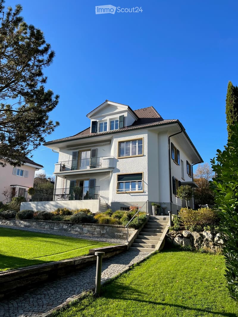 Two-story house with white exterior, multiple windows, and a balcony on the upper level. The house is surrounded by a well-maintained garden with a stone wall, steps, and greenery.