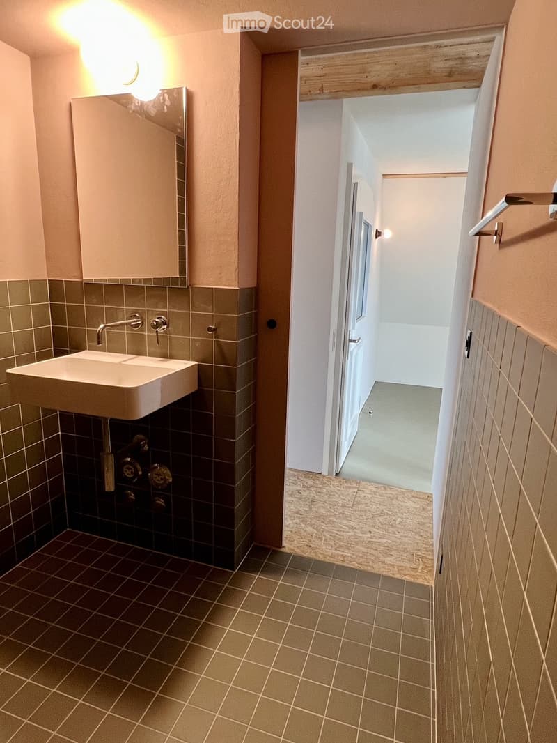 Hallway with tiled floor, beige walls, and a small sink area visible on the left side. The hallway leads to another room with a window and a door.