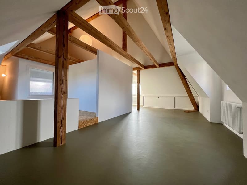 An empty attic space with exposed wooden beams, white walls, and a green floor. There are windows on the left and right sides, and a radiator on the far right wall.
