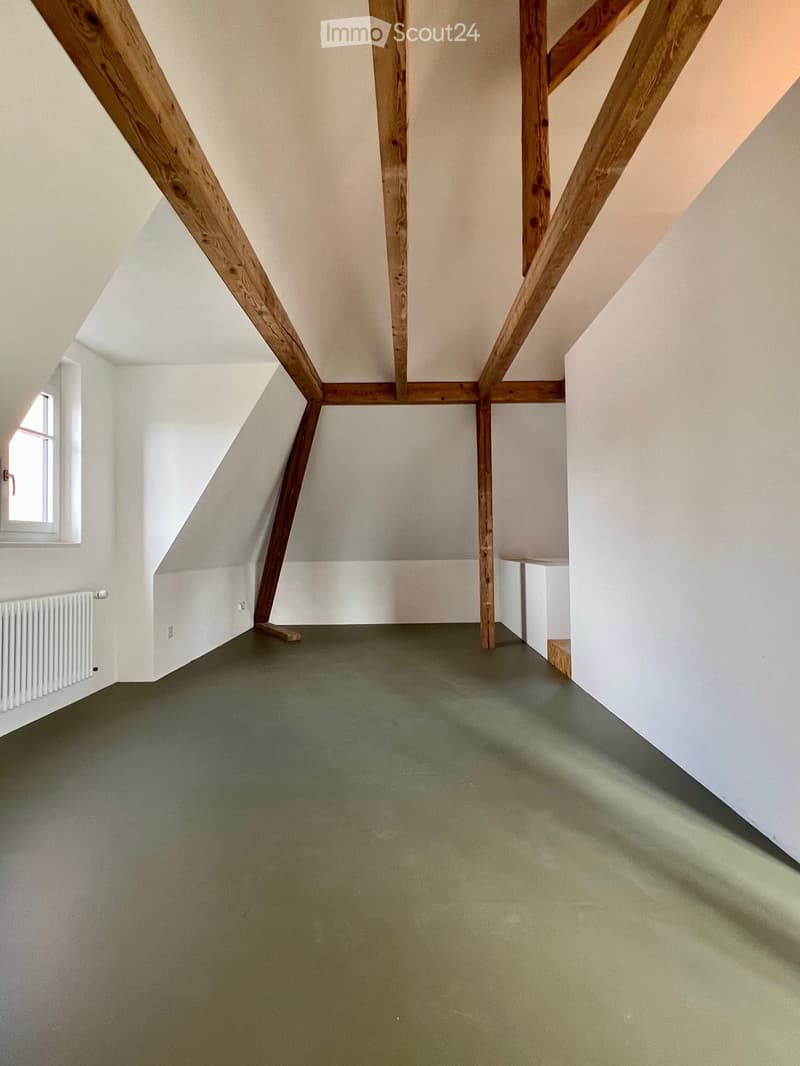 Empty attic room with exposed wooden beams, green floor, white walls, and a small window on the left side.