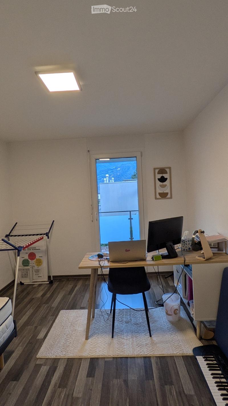 A small, well-lit room with a wooden desk, laptop, screen, and chair. A white rug is on the floor, and a window with a view of a balcony is visible.