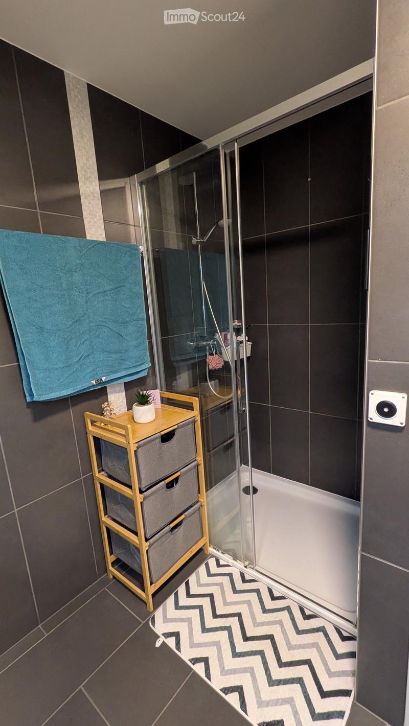 Modern bathroom with dark tiled walls and floor, glass-enclosed shower, wooden shelf with drawers, towel, and small plant.