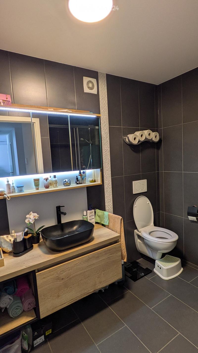 Modern bathroom with dark tiles, a black sink, a toilet, a large mirror with built-in lighting, and a wooden cabinet with towels and toiletries.