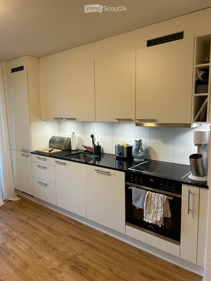 Modern kitchen with white cabinets, black countertops, and stainless steel appliances including an oven, toaster, and coffee maker. Wooden flooring and a sink with a faucet are visible.