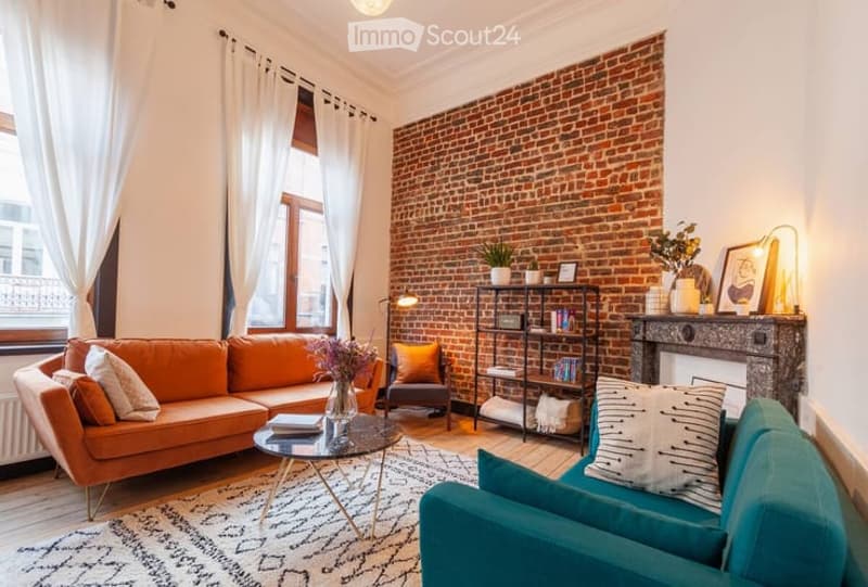 Living room with exposed brick wall, large window with white curtains, orange sofa, teal armchair, coffee table, bookshelf, fireplace, and decorative items.