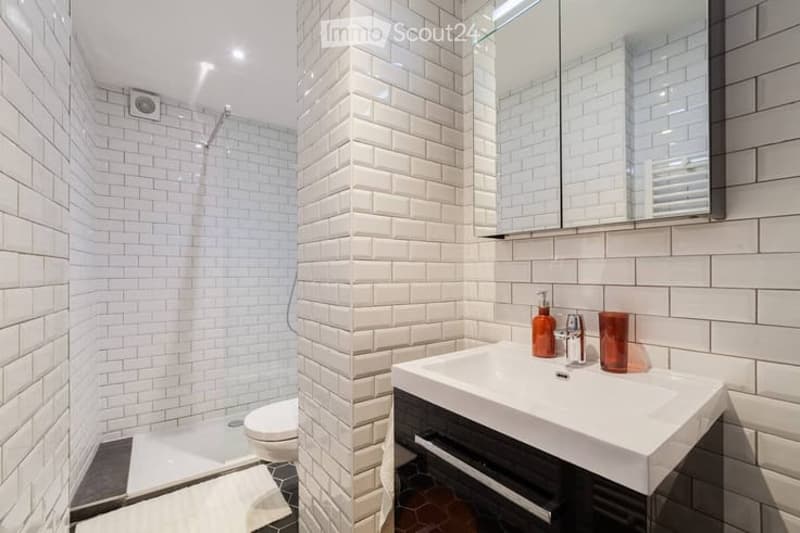 Modern bathroom with white brick tiles, a large mirror above the sink, a glass-enclosed shower, and a toilet. The sink has a sleek black cabinet and a chrome faucet.