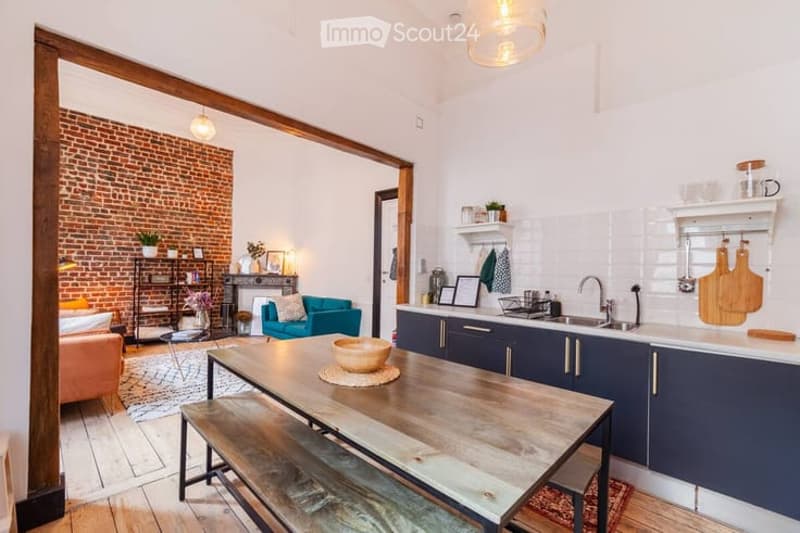 Open-plan living and dining area with exposed brick wall, modern kitchen, wooden dining table, teal armchair, and decorative shelves.