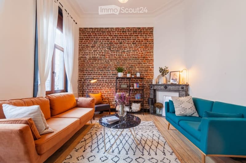 Modern living room with exposed brick wall, two sofas (orange and blue), coffee table, armchair, floor lamp, fireplace, shelf with decorative items, and vases.