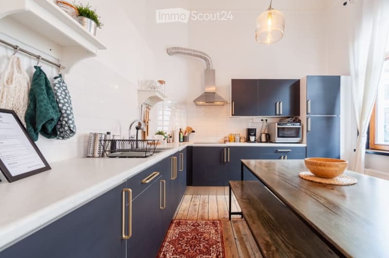 Modern kitchen with dark blue cabinets, white countertops, wooden flooring, and a small dining area with a wooden table and bowl.
