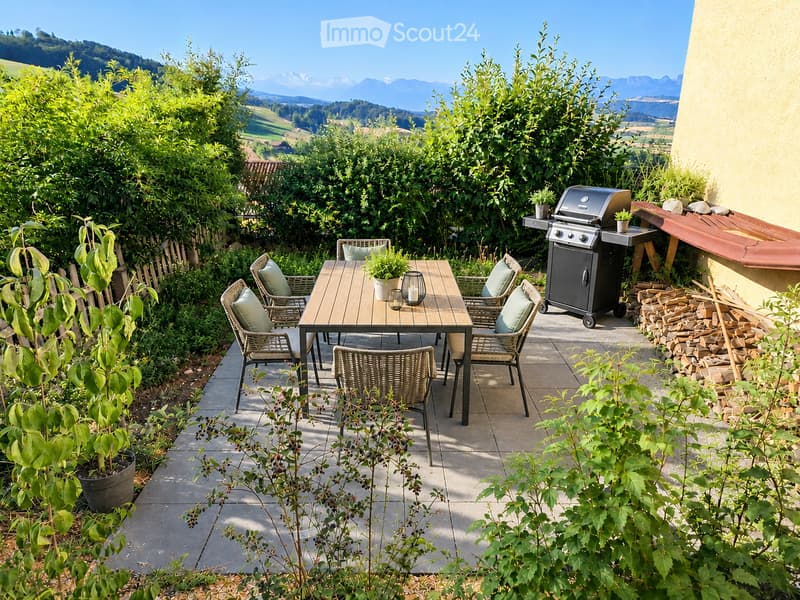 A well-maintained outdoor patio with a wooden table, six wicker chairs, a grill, potted plants, and a stack of firewood, surrounded by lush greenery and a scenic mountain view.