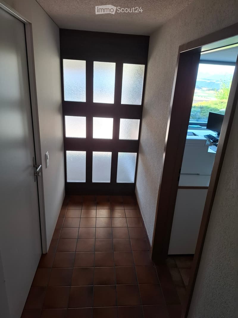 A narrow hallway with tiled flooring, a closed door with frosted glass panels, and an open doorway leading to a room with a desk and computer visible in the background.