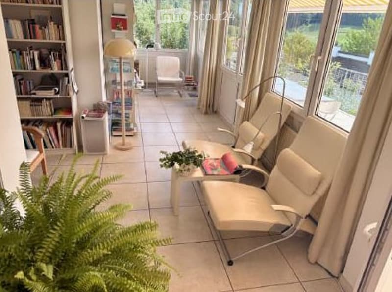 Bright living room with large windows and a view, featuring a sofa, chairs, a small table, bookshelf, and a floor lamp.