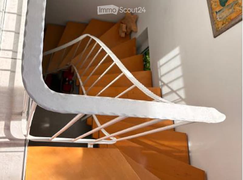 Spiral staircase with wooden steps and white handrail, leading to an upper level. The walls are white, and there is a small plant visible on the landing.