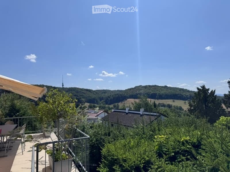 Balcony with a scenic view of a valley, trees, and houses below. Includes a small table and chairs, potted plants, and a railing.